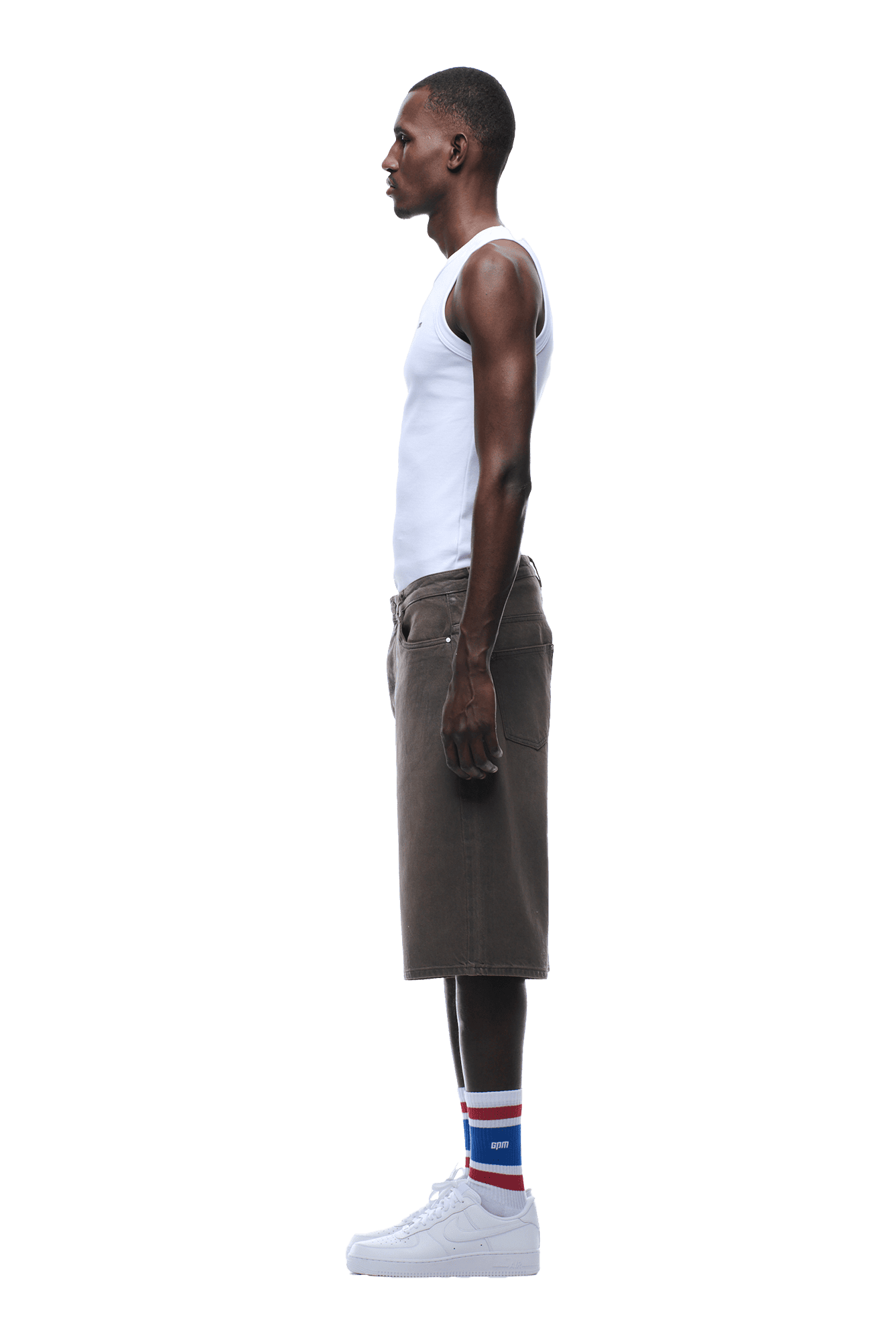 A man stands in profile, wearing a white tank top, 6PM COATED DENIM SHORTS WASHED BROWN, white sneakers, and white socks with red and blue stripes. The background is transparent.