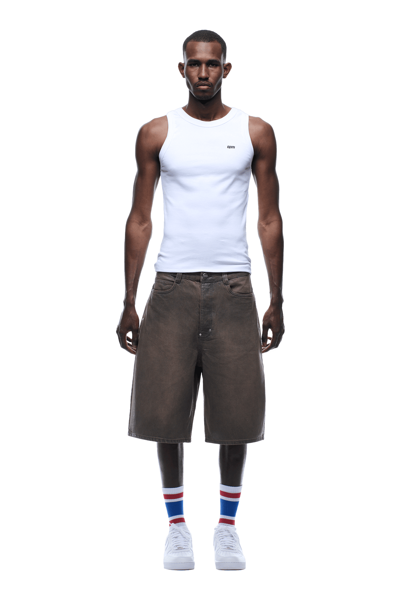 A man faces forward wearing a white tank top, COATED DENIM SHORTS WASHED BROWN by 6PM, white sneakers, and white socks with blue and red stripes. The background is transparent.