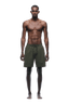 A man with a lean, muscular build stands barefoot facing forward, wearing 6PM SEERSUCKER SWIM SHORTS OLIVE. He is shirtless, arms relaxed at his sides, in front of a plain green background.