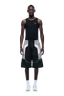A man stands facing forward, wearing a sleeveless black shirt, 6PM BAGGY WARM UP SHORTS BLACK, white socks, and white sneakers against a plain background.