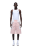 A man stands facing forward against a plain green background, wearing a fitted white tank top, 6PM BAGGY WARM UP SHORTS PINK, white socks, and white sneakers.