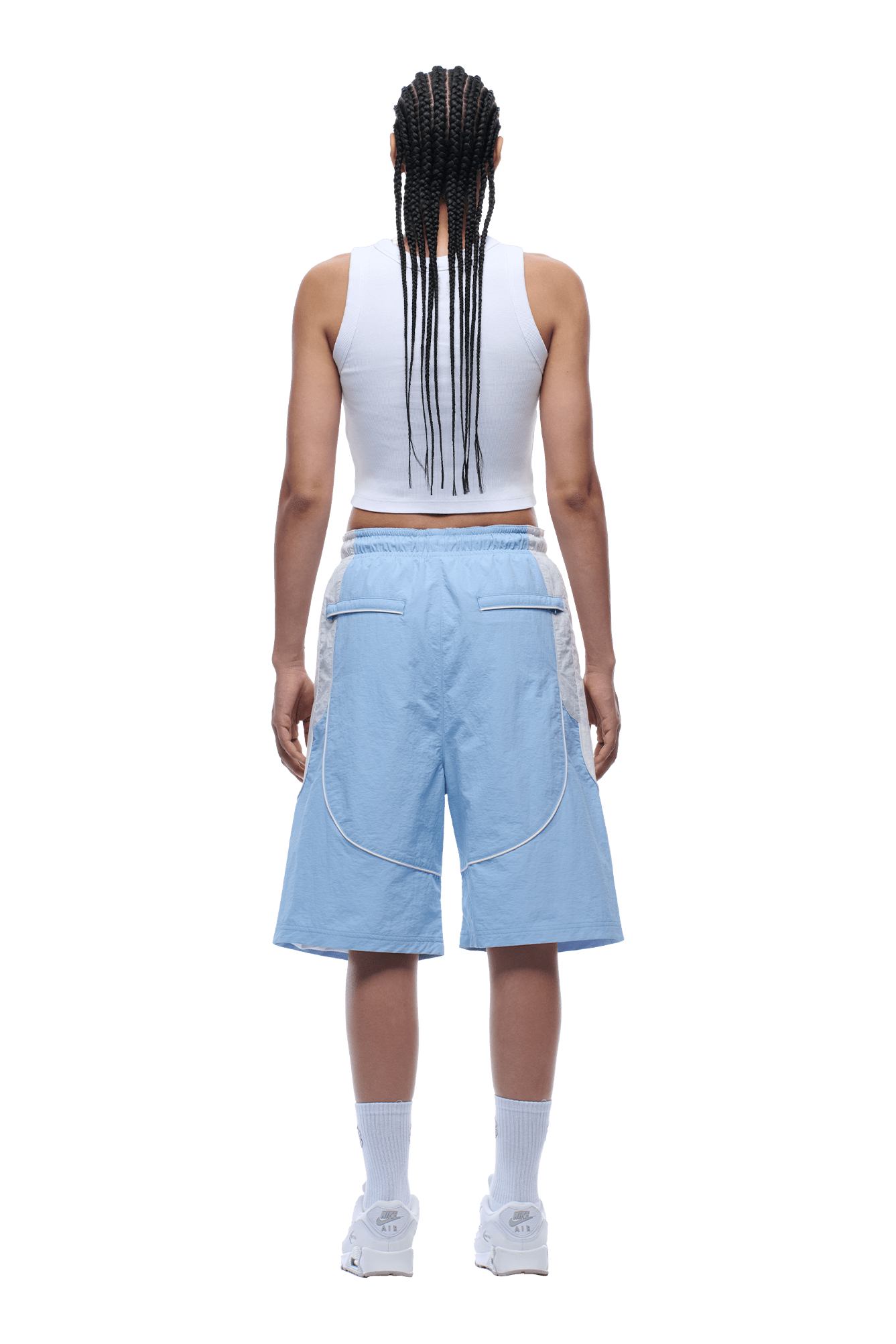 A person with long braids is shown from the back, wearing a white sleeveless crop top, 6PM BAGGY WARM UP SHORTS ICE BLUE, white crew socks, and white sneakers, standing on a plain background.