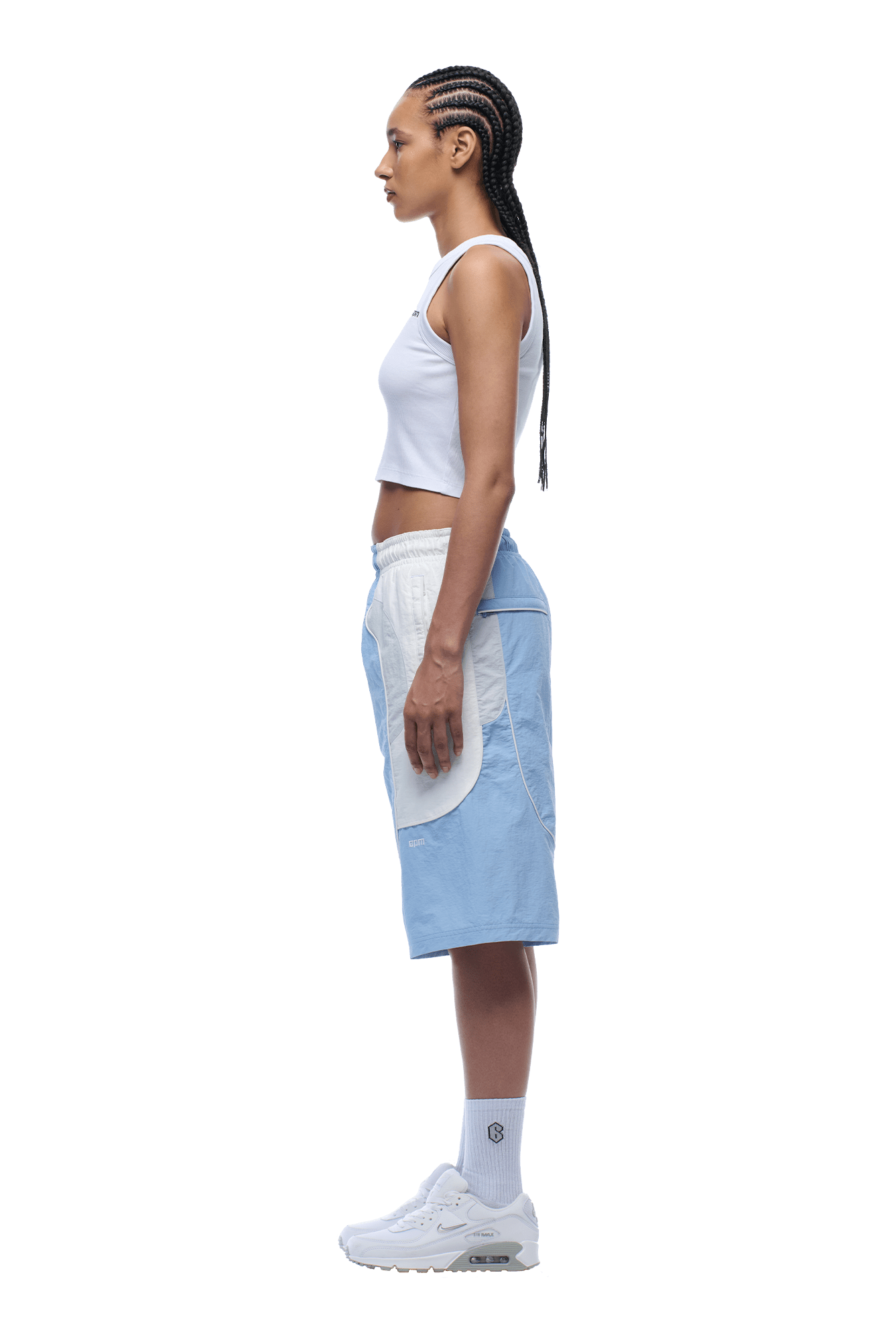 A woman with braided hair stands in profile against a plain background, wearing 6PM BAGGY WARM UP SHORTS ICE BLUE, a white crop top, white socks, and white sneakers.