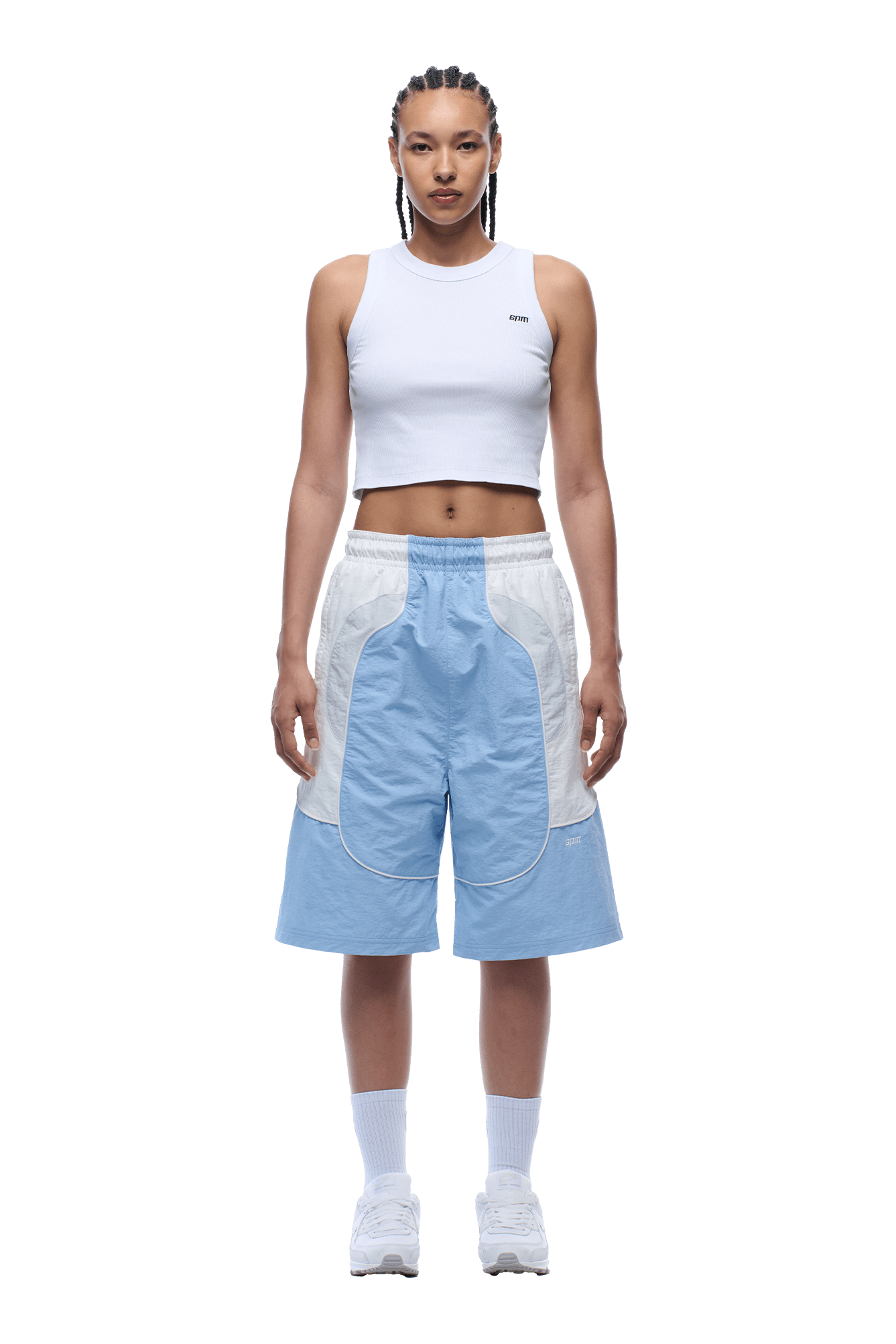A person with braided hair and a neutral expression stands facing forward, wearing a white crop top, 6PM BAGGY WARM UP SHORTS ICE BLUE, white socks, and white sneakers. The background is transparent.
