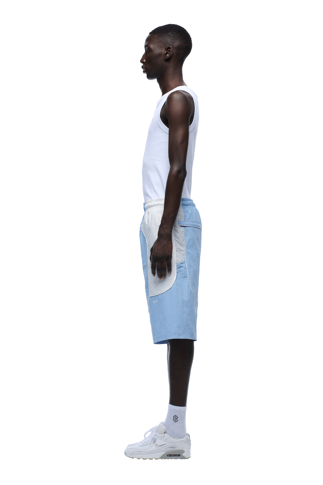A young man in profile stands against a transparent background, wearing a white tank top, 6PM BAGGY WARM UP SHORTS ICE BLUE, white socks, and white sneakers. His arms rest by his sides as he looks straight ahead.