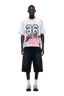 A man faces forward, wearing the 6PM CAMO JERSEY FLAMINGO—a white and pink graphic jersey with the number 26—paired with wide black knee-length shorts, white socks, and white sneakers with orange accents.