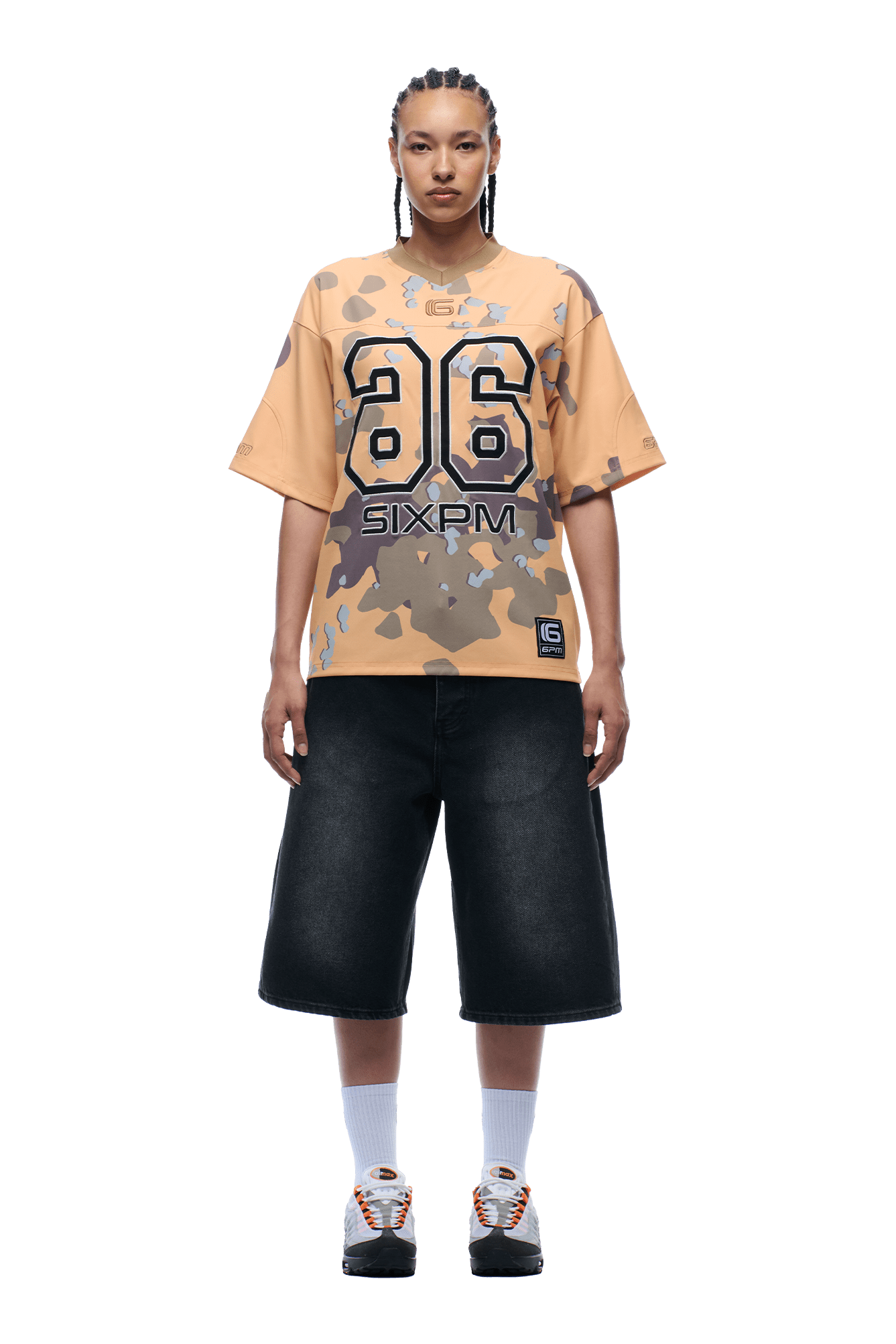 A person with braided hair wears the CAMO JERSEY BURRO shirt by 6PM, wide black knee-length shorts, white socks, and sneakers, standing facing forward against a transparent background.