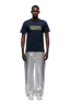 A man with short hair stands facing forward in a 6PM CLEAN EDGE WAVE T-SHIRT MIDNIGHT BLUE, featuring yellow text, paired with wide metallic silver pants and white sneakers, set against a plain white background.