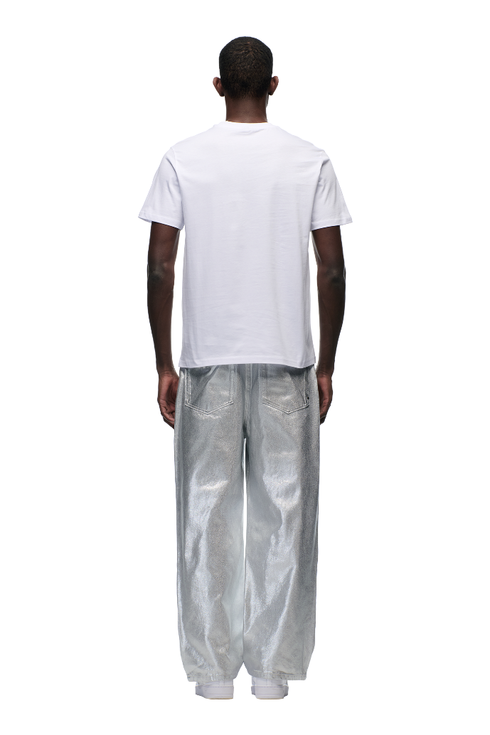 A person stands facing away in the 6PM DOWN UNDER COLLEGE T-SHIRT WHITE, which features a woven side label, paired with loose silver pants against a plain white background.