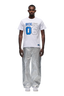 A man stands facing forward wearing the 6PM DOWN UNDER COLLEGE T-SHIRT in white, paired with shiny silver wide-leg pants and white sneakers, set against a plain white background.