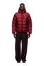 A person faces forward in a 6PM LIGHT PUFFER JACKET RECTANGLES WINE RED with an adjustable hood and zippered chest pocket, paired with loose dark brown pants and black shoes against a plain white background for a cozy sports-inspired look.