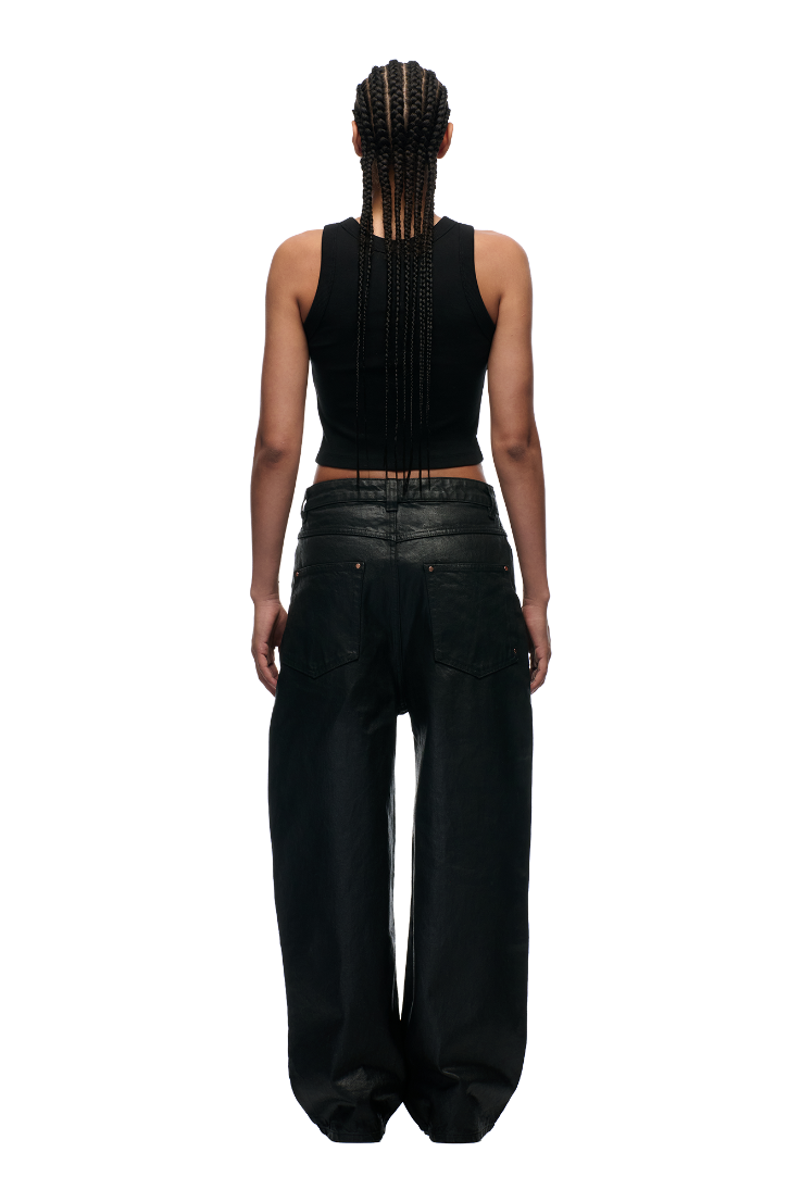 A person with long braids, dressed in a black sleeveless crop top and 6PM WAX COATED DENIM BLACK wide jeans with metal buttons, stands facing away against a plain white background.