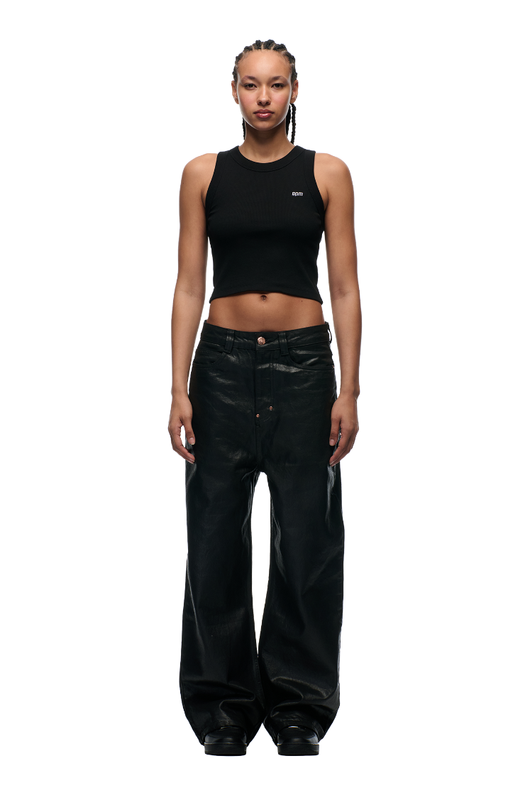 A person with braided hair models the WAX COATED DENIM BLACK pants by 6PM, featuring metal buttons, paired with a fitted black crop tank, standing against a plain white background and facing forward with a neutral expression.