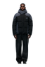 A man faces forward against a white background, wearing 6PM's COLOUR BLOCKING PUFFER BLACK/ANTHRACITE with a removable hood, wide-leg black pants, and black shoes.