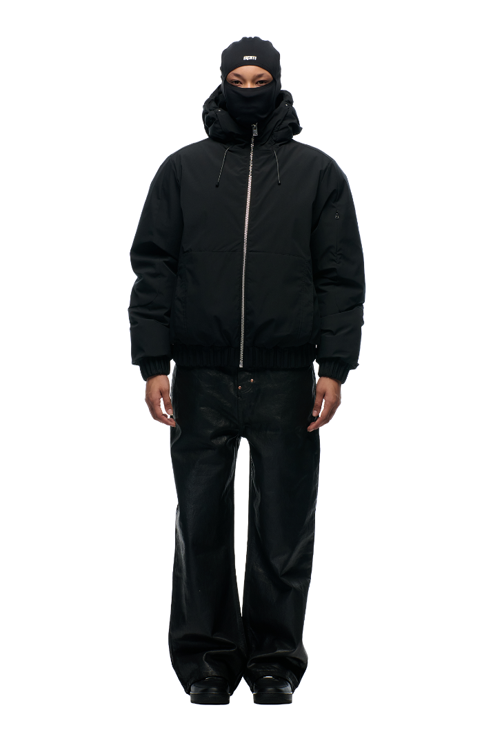 A person stands facing forward against a light background wearing the 6PM EMBOSSED JACKET BLACK with a hidden arm pocket, black balaclava, loose black jeans, and black shoes.