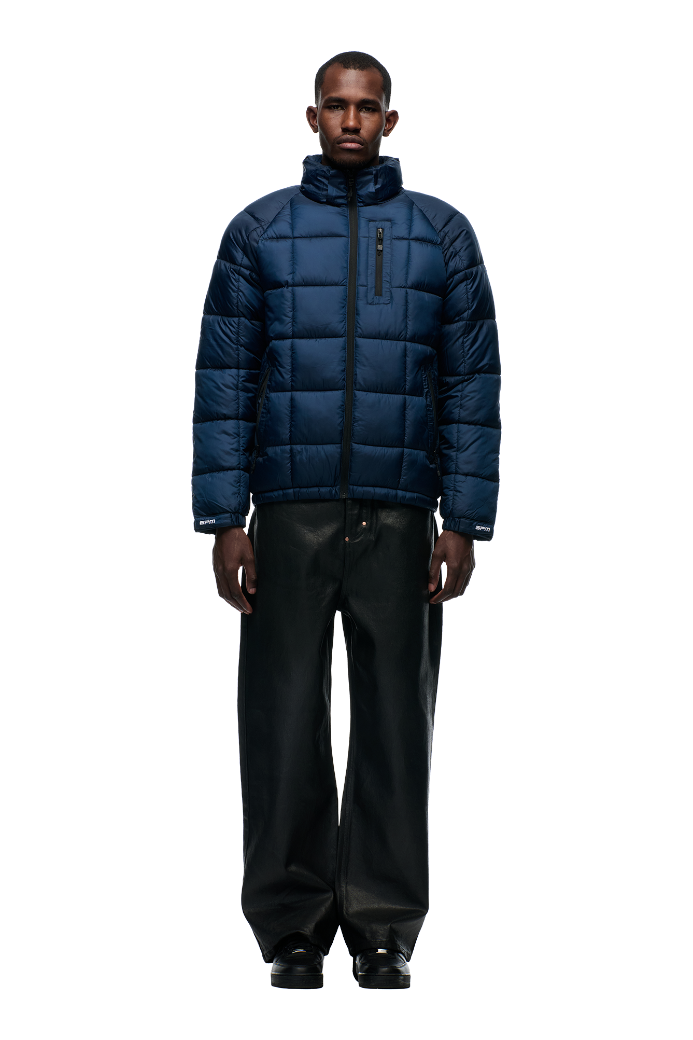 A man faces forward in a 6PM LIGHT PUFFER JACKET RECTANGLES MIDNIGHT BLUE with sports embroidery, a zip-up front, and black loose-fitting pants, set against a plain white background.