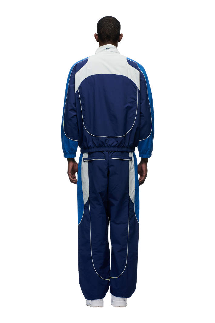 A person stands facing away, wearing the 6PM EVERYDAY TRACK JACKET SHADOW BLUE with embroidered logos and matching pants, set against a plain white background.