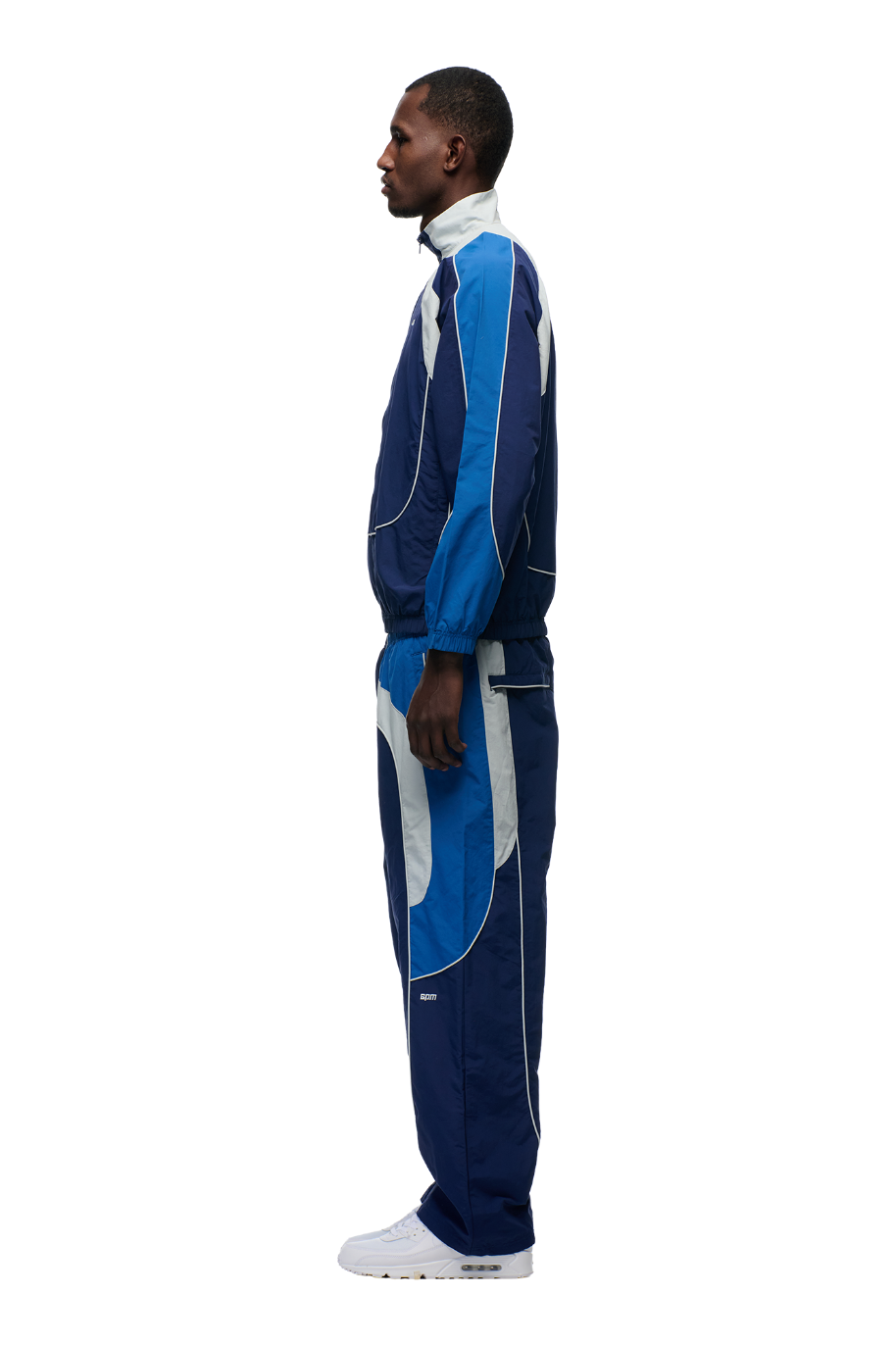 A man in profile wears the 6PM EVERYDAY TRACK JACKET SHADOW BLUE—featuring blue and white color-blocked nylon, mesh lining, and subtle embroidered logos—paired with white sneakers against a plain white background.