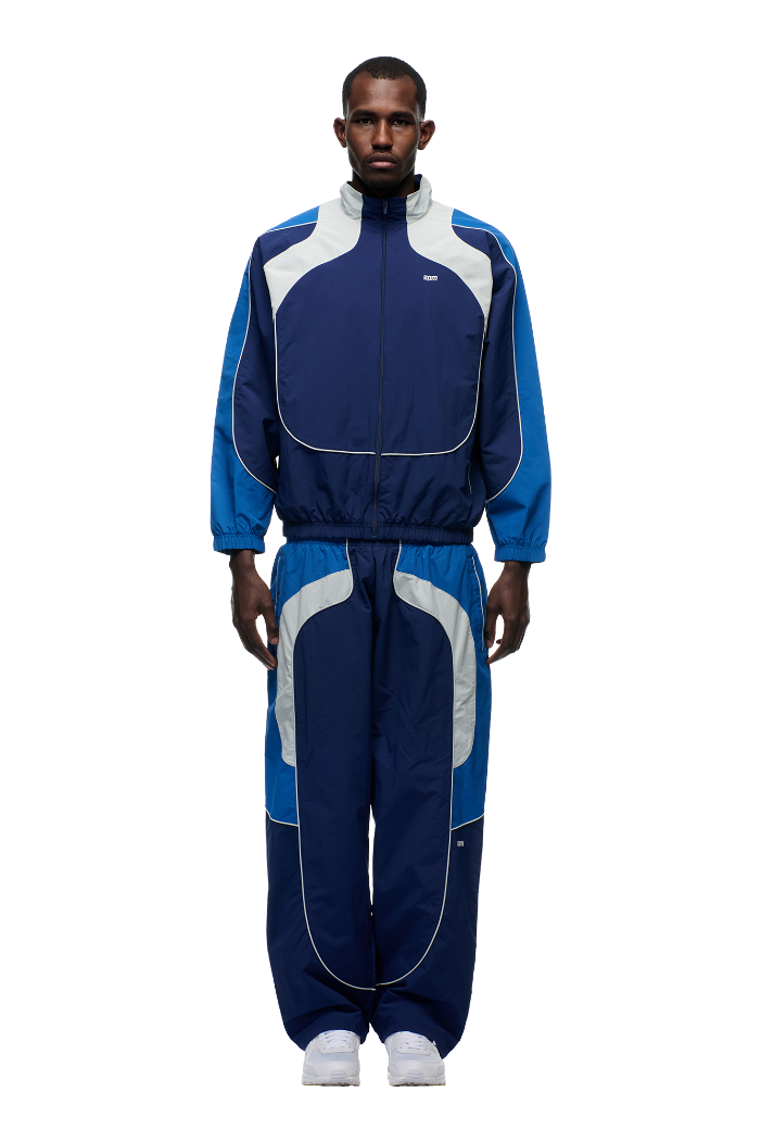 A man stands facing forward, wearing the 6PM EVERYDAY TRACK JACKET SHADOW BLUE with matching track pants and white sneakers, against a plain white background.