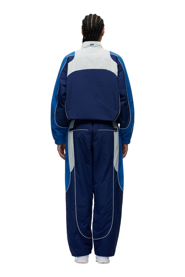 A person with braided hair, seen from behind against a plain white background, wears the 6PM EVERYDAY TRACK JACKET SHADOW BLUE featuring color-blocked navy, light gray, and royal blue nylon.