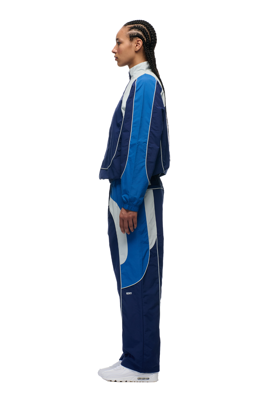 A person with tight cornrows is shown in profile, wearing the 6PM EVERYDAY TRACK JACKET SHADOW BLUE with embroidered logos and white sneakers, standing against a plain white background.