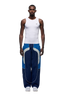 A man faces forward against a plain white background, wearing a fitted white tank top, white sneakers, and 6PM’s EVERYDAY TRACK PANTS SHADOW BLUE with white and gray curved accents and zipped pockets.