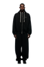 A man stands facing forward against a plain light background, wearing the 6PM BOXY ZIP HOODIE BLACK with white drawstrings, embroidered logo, metal zipper, and matching wide-leg black pants and black shoes.