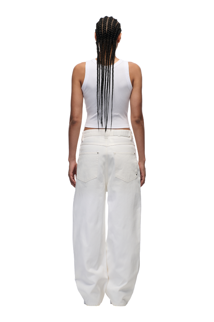 A person with long braids stands with their back to the camera, wearing a white sleeveless top and 6PM's WAX COATED DENIM WHITE pants against a plain white background.