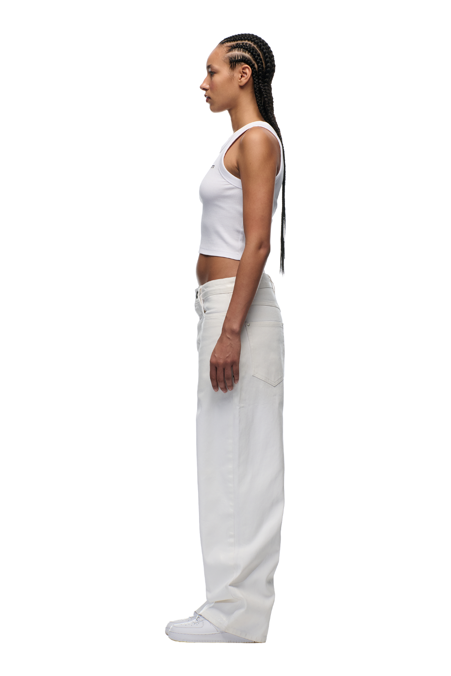 A person with braided hair stands in profile, wearing a white sleeveless crop top, 6PM's WAX COATED DENIM WHITE pants with a leather badge and branded buttons, and white shoes against a plain light background.