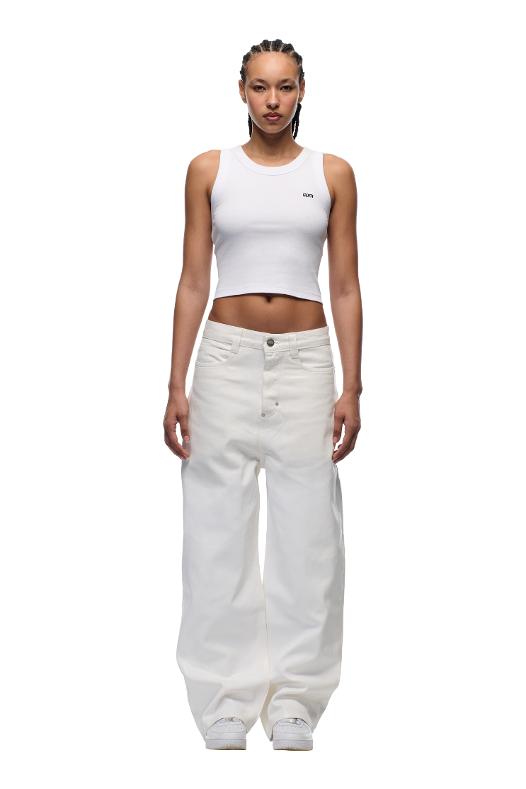 A woman with braided hair models the 6PM WAX COATED DENIM WHITE pants and a fitted white crop tank top, paired with white sneakers. She stands against a plain white background, with subtle leather badge details finishing her look.