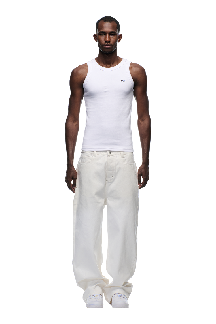 A man stands facing forward against a plain white background, wearing 6PM's WAX COATED DENIM WHITE jeans, a fitted white tank top, and white sneakers.