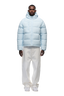 A man stands facing forward in a 6PM PUFFER JACKET ICE BLUE with an adjustable hood, paired with white baggy pants and white sneakers, against a plain white background.
