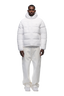 A man faces forward, wearing the 6PM PUFFER JACKET WHITE with an adjustable hood, paired with loose white pants and white sneakers, set against a plain light background.