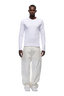 A man stands facing forward against a plain white background, wearing the 6PM 6PM+ RIBBED LS WHITE fitted long-sleeve top, loose white pants, and white sneakers.