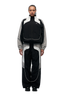 A person with braided hair faces forward, wearing the 6PM EVERYDAY TRACK JACKET BLACK featuring white piping, embroidered logos, mesh lining, and black shoes, set against a plain light background.