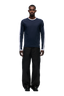 A man stands facing forward against a light gray background, wearing the 6PM GRAFFITI RINGER LS MIDNIGHT BLUE top, loose black pants, and black shoes.