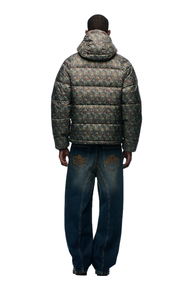 A person stands facing away against a white background, wearing the 6PM PUFFER JACKET MINI CAMO—a water-repellent puffer with adjustable hood and mini camo print—paired with loose blue jeans featuring embroidered back pockets.