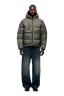 A man stands facing forward, dressed in a 6PM PUFFER JACKET MINI CAMO with an adjustable hood, wide-leg faded blue jeans, and black shoes against a plain white background.