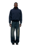 A man faces forward in a 6PM COLLEGE LETTER HOODIE MIDNIGHT BLUE, featuring back embroidery and 6PM lettering, paired with loose, faded blue jeans and black shoes, set against a plain light background.