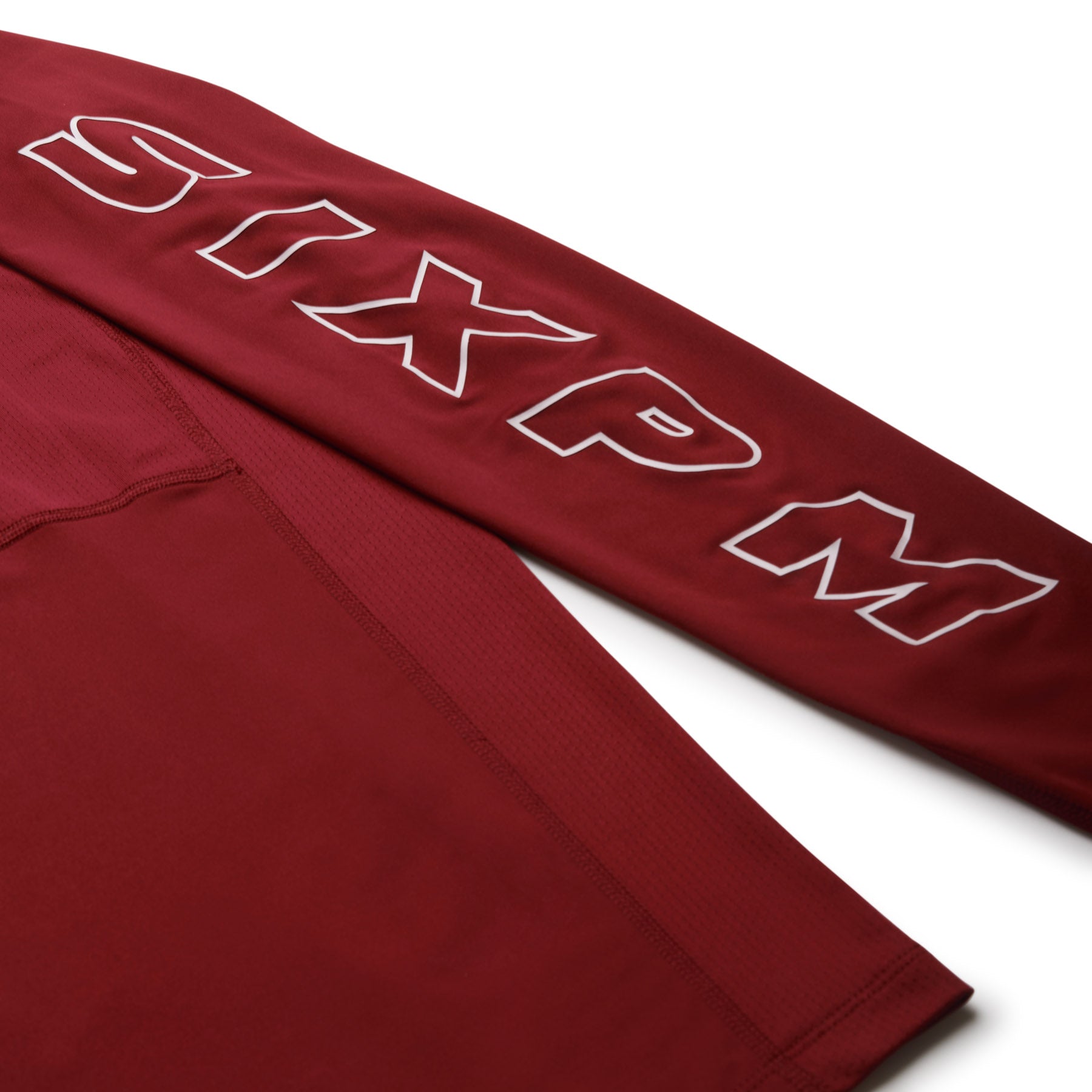 A close-up of the 6PM SPORTS LONGSLEEVE WINE RED by 6PM shows “SIXPM” printed in bold, outlined letters along the sleeve on smooth, slightly textured fabric.