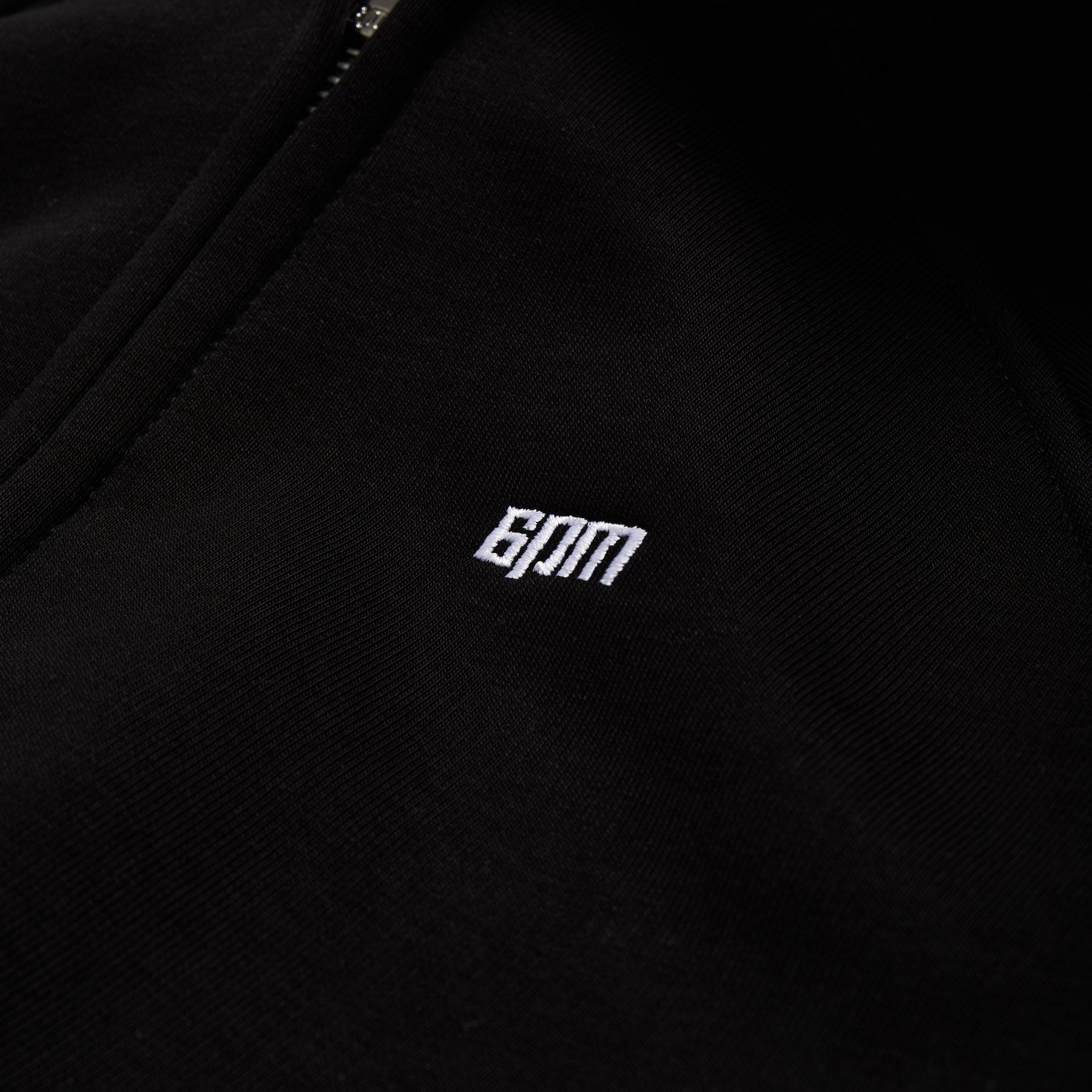 Close-up of black fabric from the 6PM WMNS CROPPED ZIP HOODIE BLACK, featuring a small white embroidered 6PM logo.