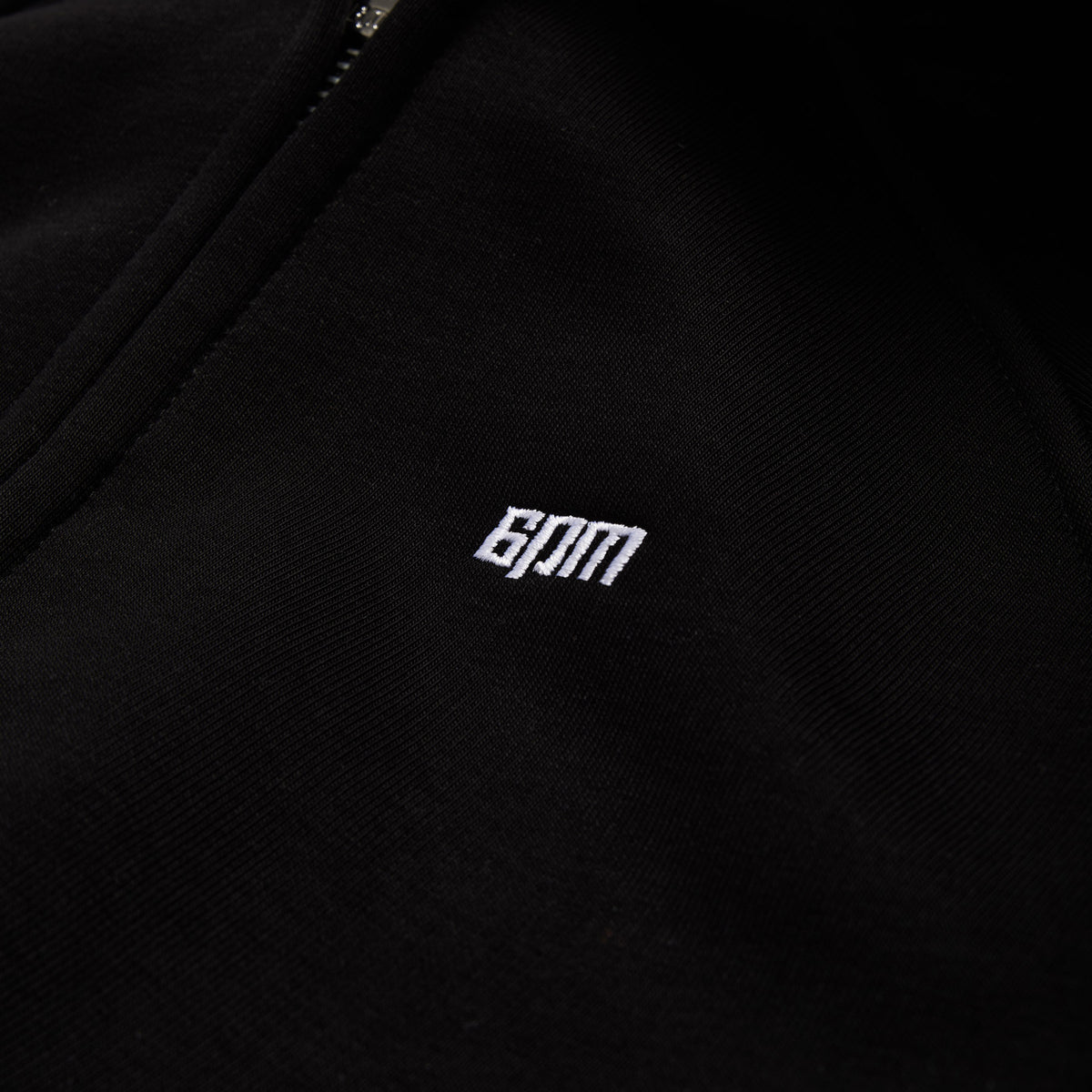 Close-up of black fabric from the 6PM WMNS CROPPED ZIP HOODIE BLACK, featuring a small white embroidered 6PM logo.