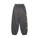 The 6PM SUPER WIDE JOGGER GREY CAMO features dark gray camo sweatpants with an elastic waistband and cuffs, and bold yellow-green varsity-style letters running vertically down the left leg, shown on a white background.