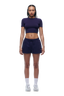A woman with braided hair faces forward, wearing the 6PM WMNS CROPPED T-SHIRT ORBIT BLUE, navy athletic shorts, white socks, and sneakers against a white background.