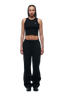 A woman with braided hair stands facing forward against a white background, wearing 6PMs WMNS OPEN LEG BLACK sweatpants, a black crop tank top, and black shoes. Her arms rest by her sides.