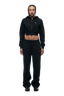 A woman with braided hair and a confident expression stands facing forward against a white background, wearing the 6PM WMNS CROPPED ZIP HOODIE BLACK, paired with black sweatpants and black sneakers.