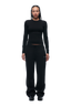 A woman with braided hair faces forward against a white background, wearing the 6PM WMNS LONGSLEEVE BLACK shirt, paired with loose black sweatpants and black sneakers.