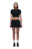 A woman with braided hair faces forward against a white background, wearing the 6PM WMNS CROPPED T-SHIRT BLACK, black shorts, white crew socks, and black sneakers.