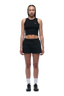 A woman with braided hair and a neutral expression faces forward against a plain white background, wearing 6PM’s WMNS SHORTS BLACK, a black sleeveless crop top, white socks, and black athletic shoes.