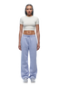 A woman with braided hair and a neutral expression stands against a white background, wearing the 6PM WMNS CROPPED T-SHIRT OFF-WHITE, paired with light blue sweatpants and white sneakers.